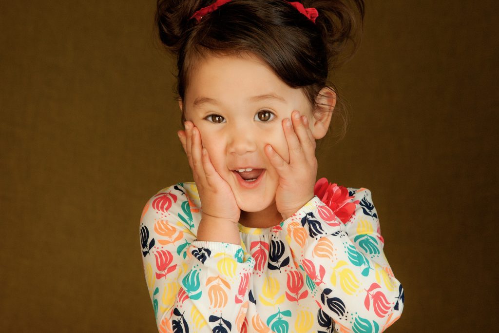 Young child with dark hair in buns wearing colorful floral shirt, hands on cheeks, surprised expression against tan background.