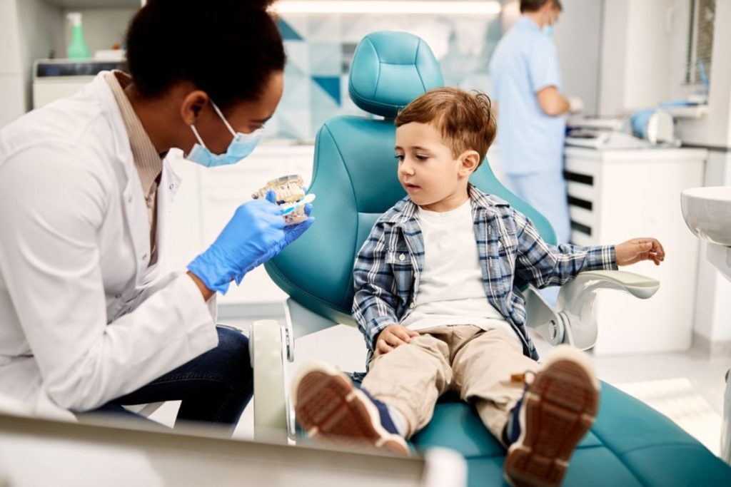 Pediatric dentist showing dental model to young boy in clinical examination chair.