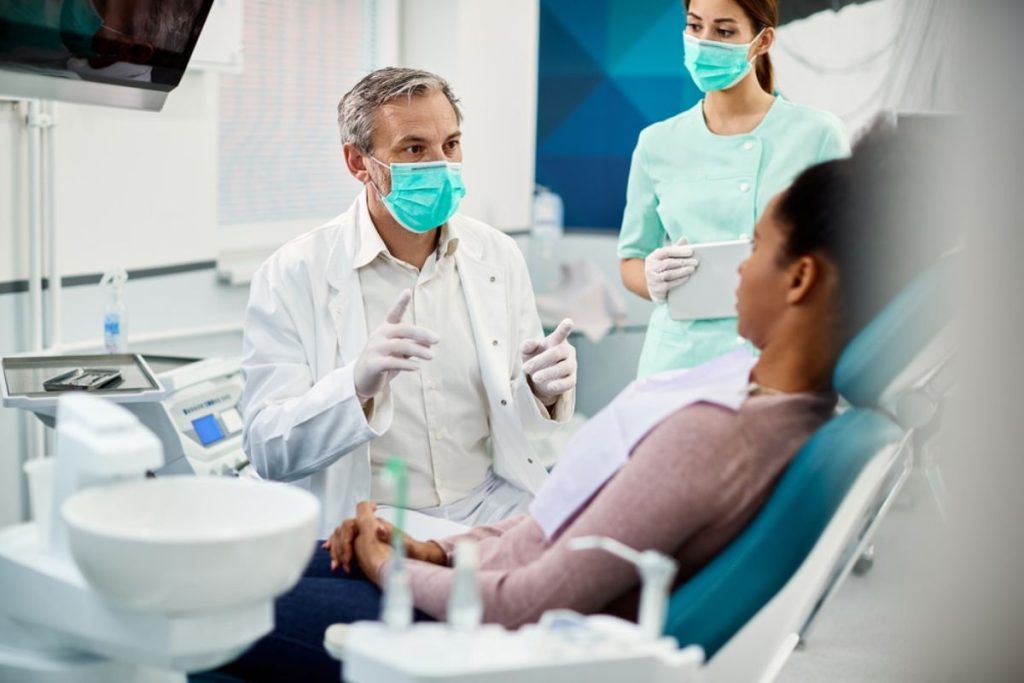 Dentist in white coat and mask explains treatment to pregnant patient in dental chair with assistant present.