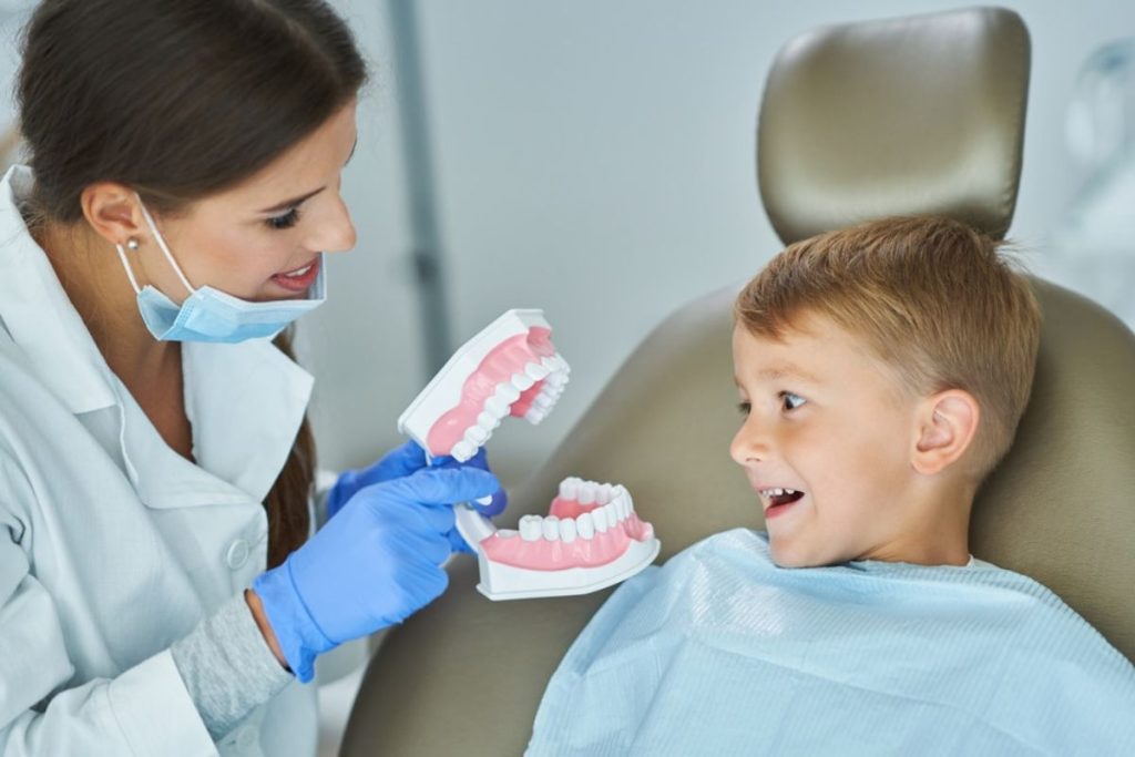 Pediatric dentist showing dental models to young boy in treatment chair during examination.