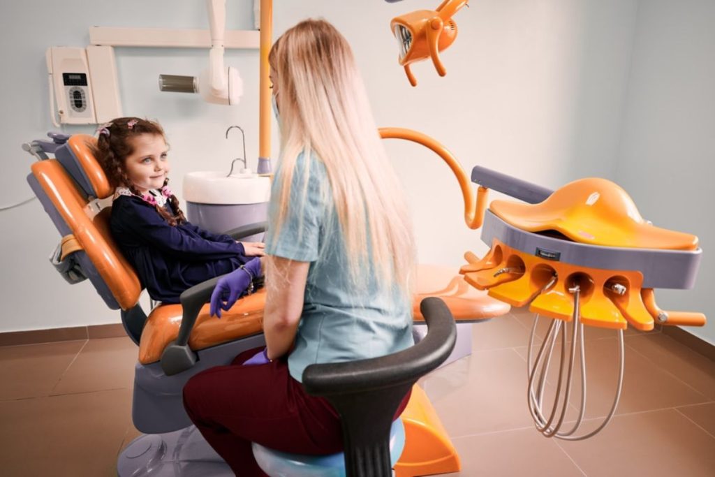 Pediatric dentist examines young girl in modern dental office with orange equipment.