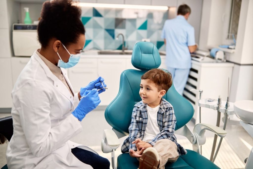 Pediatric dentist in mask and gloves showing child dental tools in modern clinic examination room.