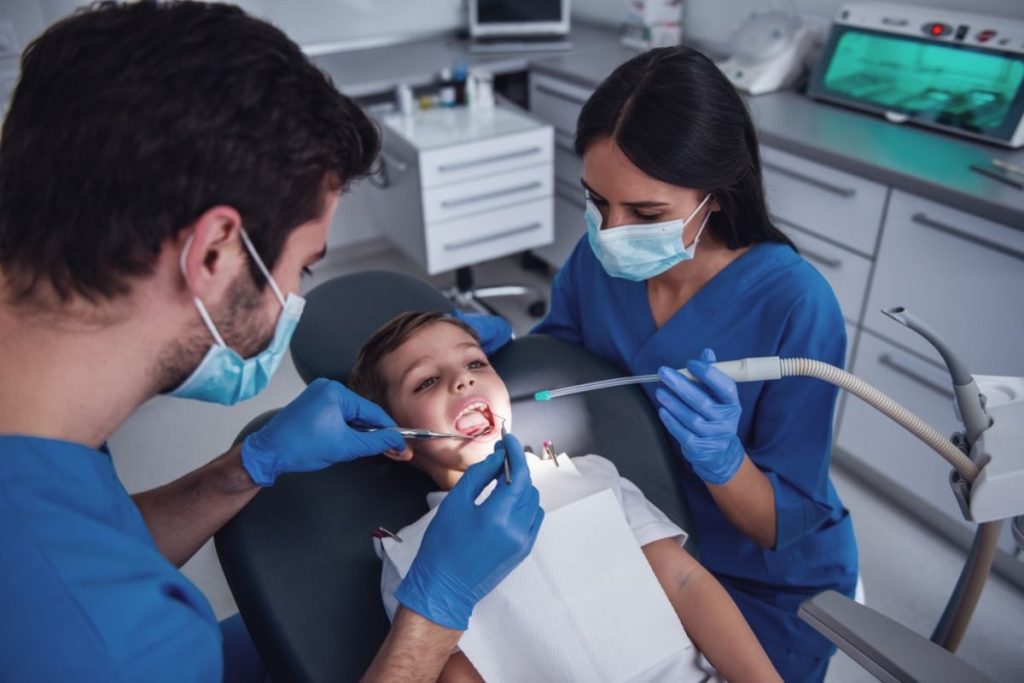 Two dentists in blue scrubs examining child's teeth in modern dental office with equipment.