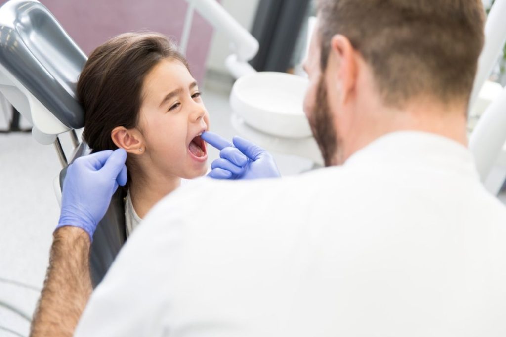Dentist in blue gloves examines young girl's mouth in dental chair during checkup.