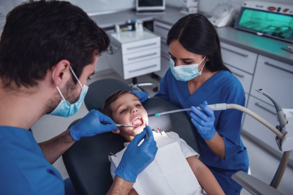Two dental professionals examine a young child's teeth in a modern dental clinic using specialized equipment.