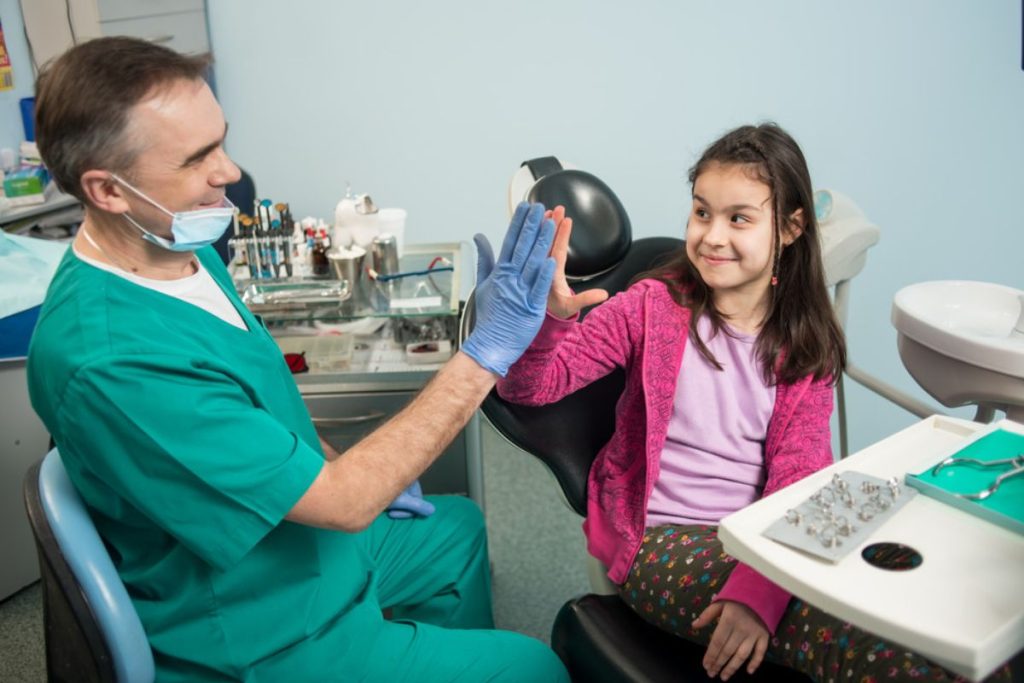 Dentist in green scrubs and mask giving high-five to smiling young girl patient in dental office.