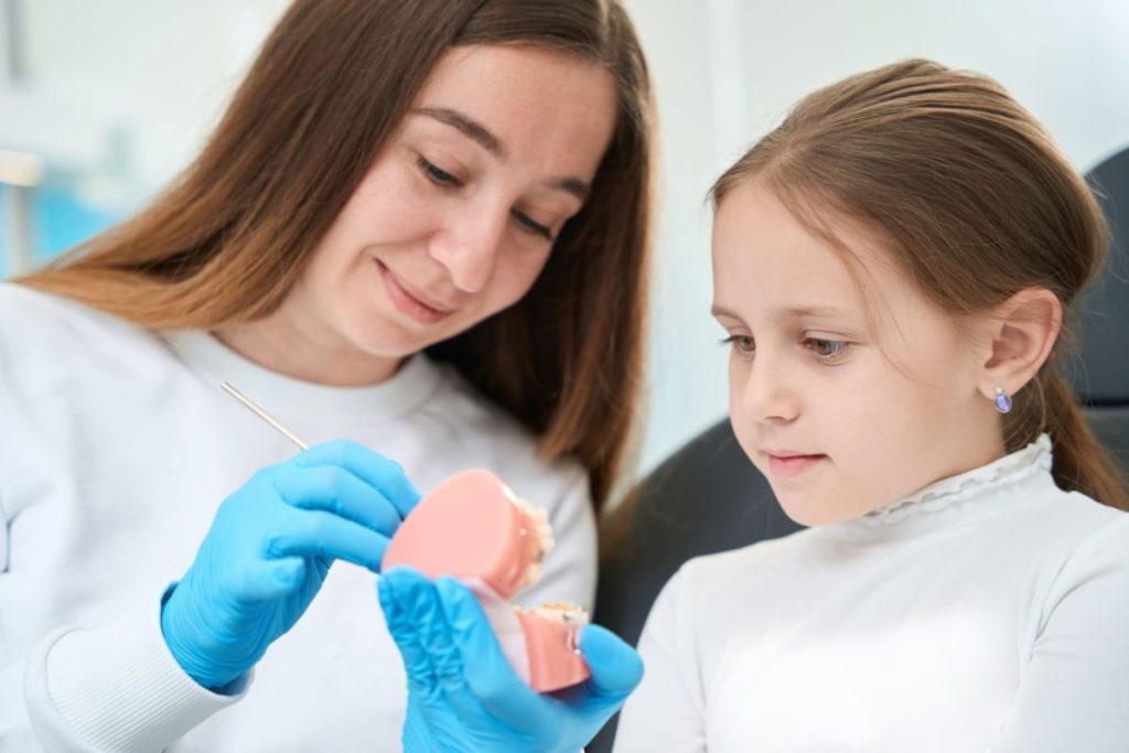 Dentist in blue gloves demonstrating dental model to young girl patient in clinical setting.
