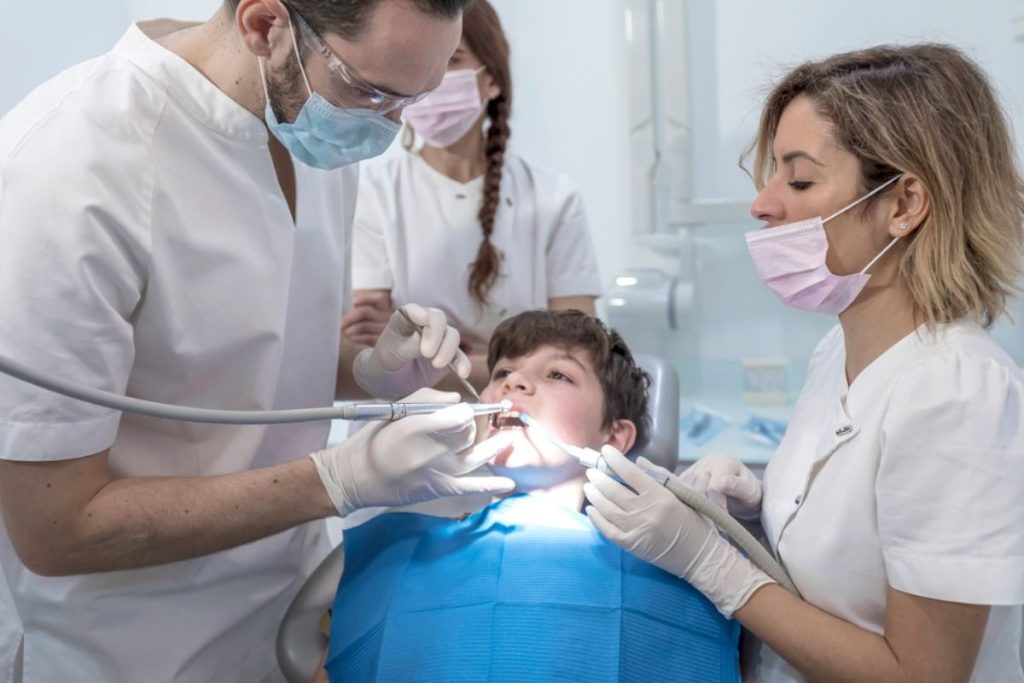 Pediatric dentist examining child's teeth with assistant and parent in dental clinic.