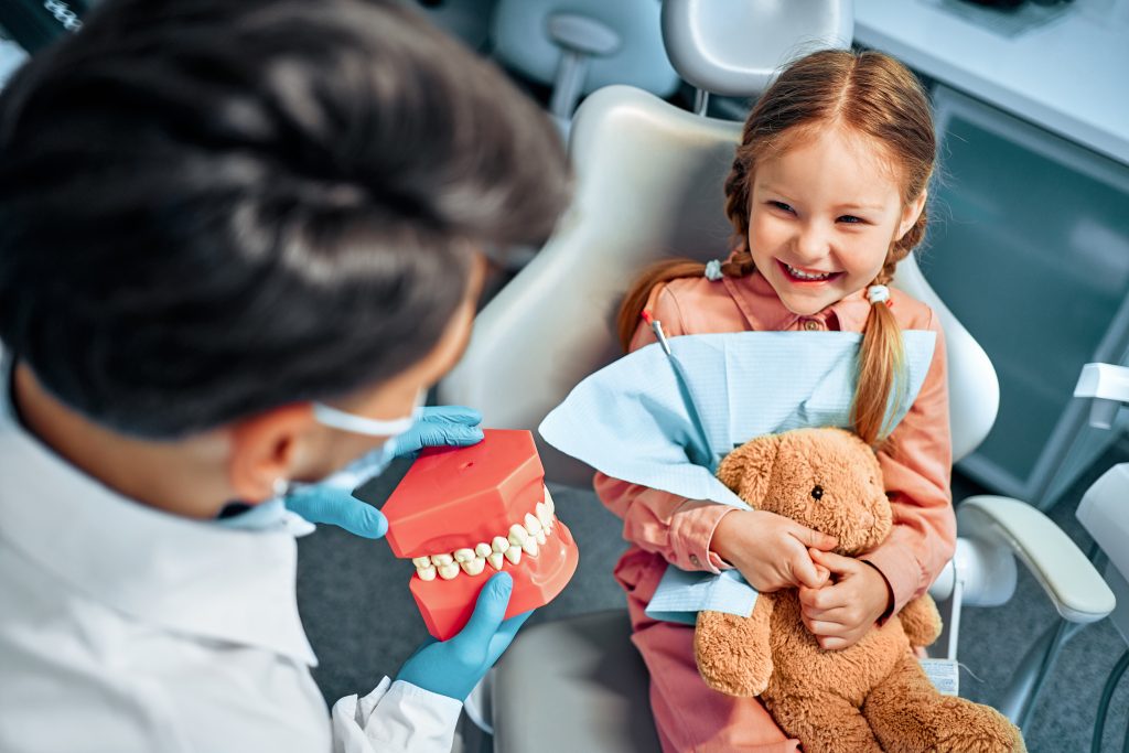 Pediatric dentist showing young girl tooth model in dental chair with teddy bear.